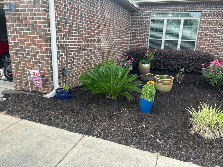 Front entrance garden bed with potted plants, shrubs, and decorative planters against a brick house with blue-trimmed window